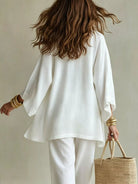 NEWYORKLISTED STYLE COLLECTION: A woman shown from behind wearing a loose white long-sleeve top and white pants, holding a woven straw tote bag against a plain beige background.