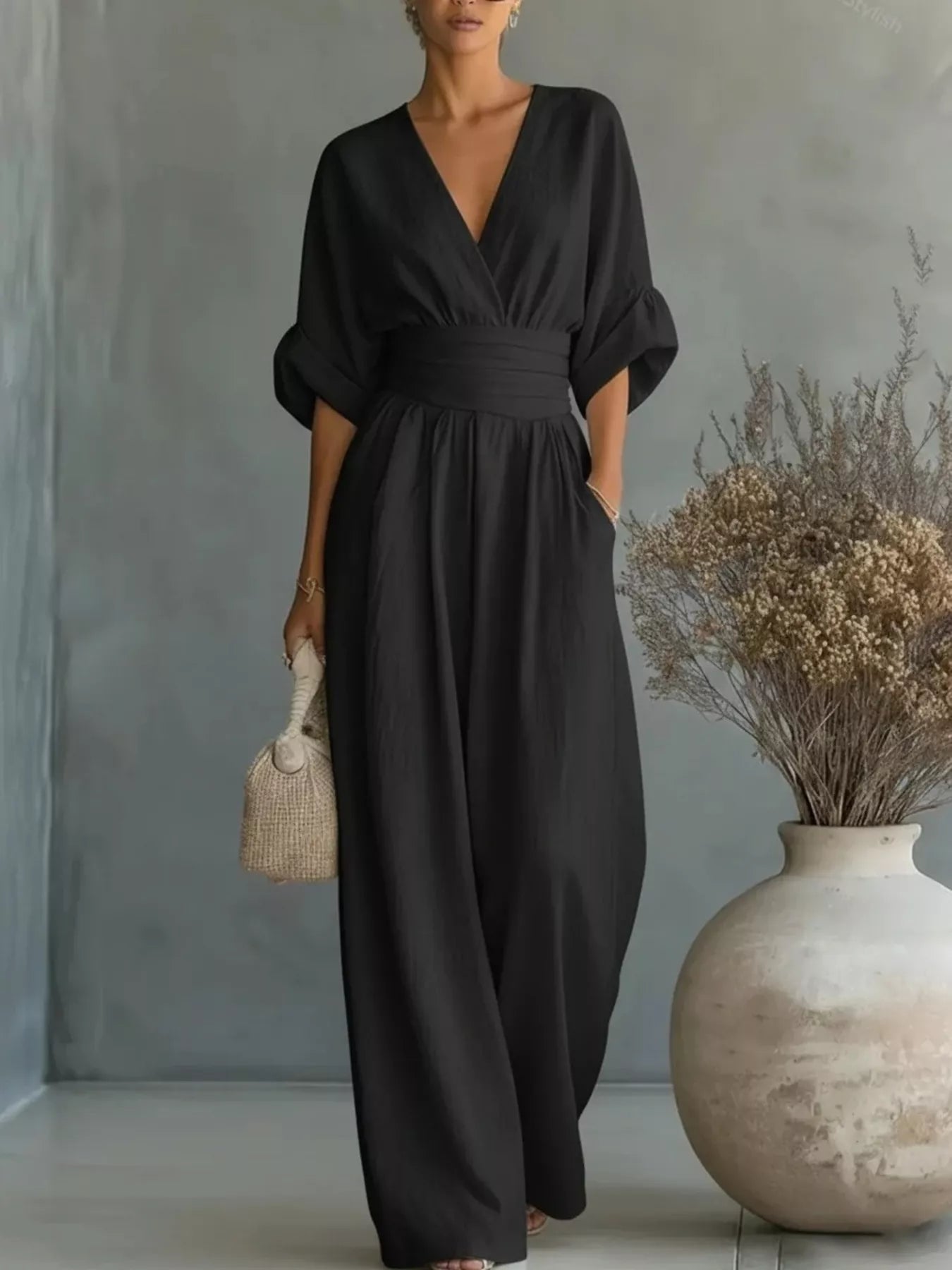 NEWYORKLISTED STYLE COLLECTION: Woman wearing a black jumpsuit standing next to a vase with dried plants against a gray wall.