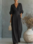 NEWYORKLISTED STYLE COLLECTION: Woman wearing a black jumpsuit standing next to a vase with dried plants against a gray wall.