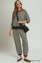 NEWYORKLISTED COMFORT COLLECTION: A woman modeling a matching black and ivory striped lounge set, featuring a long-sleeve top and jogger pants, styled with a black crossbody bag and black sneakers against a light gray studio background.