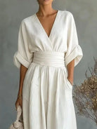 NEWYORKLISTED STYLE COLLECTION: A woman wearing a cream-colored linen jumpsuit with a deep V-neckline, wide three-quarter sleeves, and a matching fabric waist belt. She is holding a woven clutch against a textured gray background with dried floral decor.