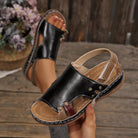 NEWYORKLISTED SHOE COLLECTION: A person holds a black leather slide sandal with a tan footbed and ankle strap, while another matching sandal is worn on a foot, set against a rustic wooden background.