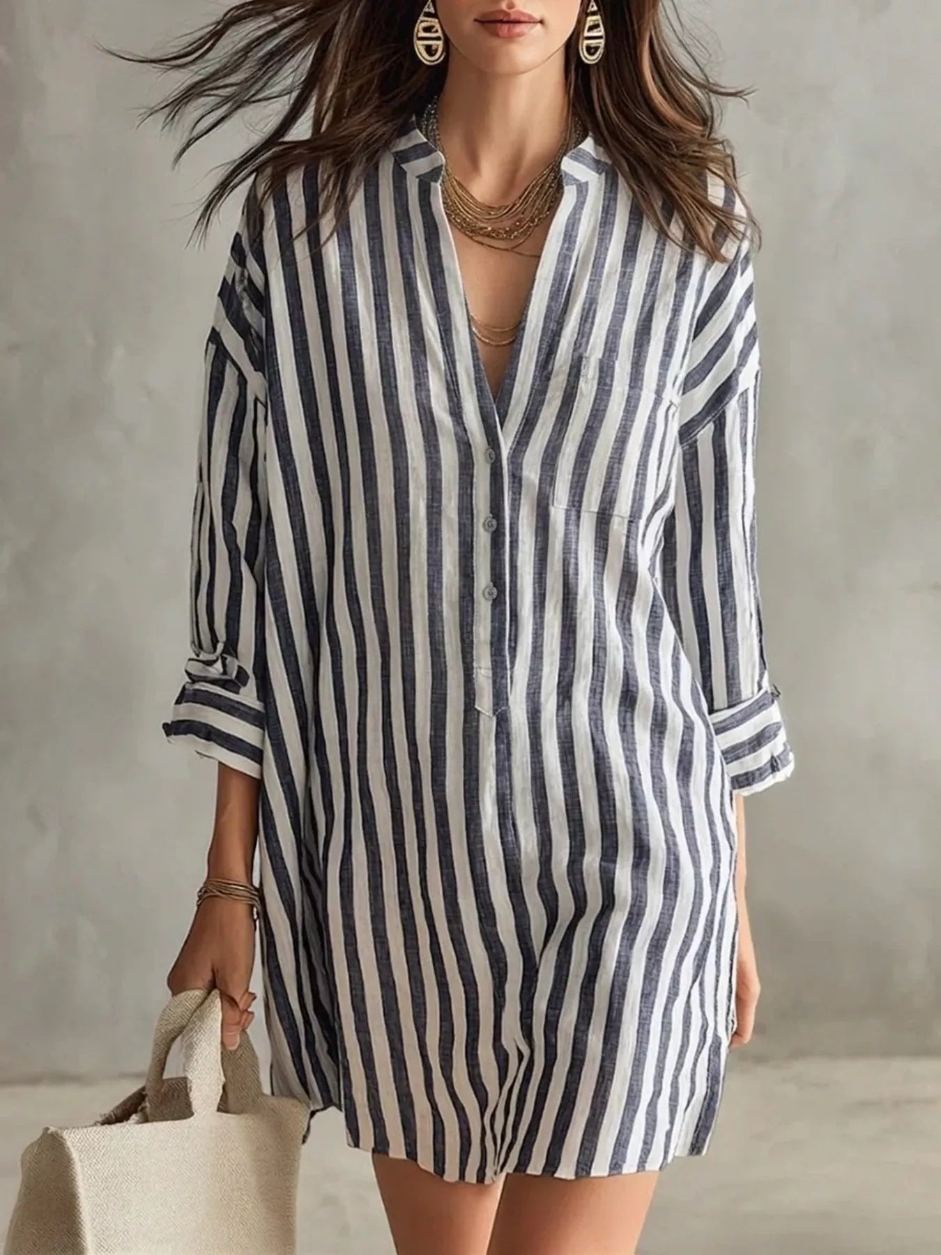 NEWYORKLISTED STYLE COLLECTION: A woman wearing a loose-fitting, short button-front shirt dress with vertical navy and off-white stripes. She is accessorized with layered gold necklaces, earrings, and bracelets, and is holding a light beige tote bag against a textured gray wall background.