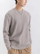 NEWYORKLISTED ATLAS MEN'S COLLECTION: A person wearing a light gray ribbed knit crewneck sweater, paired with tan pants and a silver necklace, photographed against a plain light gray background.