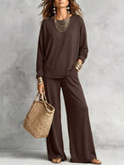 NEWYORKLISTED STYLE COLLECTION: A woman models a dark brown two-piece lounge set consisting of a long-sleeve top and matching wide-leg pants, styled with layered gold necklaces, bracelets, and a straw tote bag against a textured gray wall.