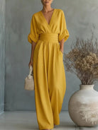 NEWYORKLISTED STYLE COLLECTION: Woman wearing a yellow jumpsuit standing next to a vase with dried flowers against a gray wall.