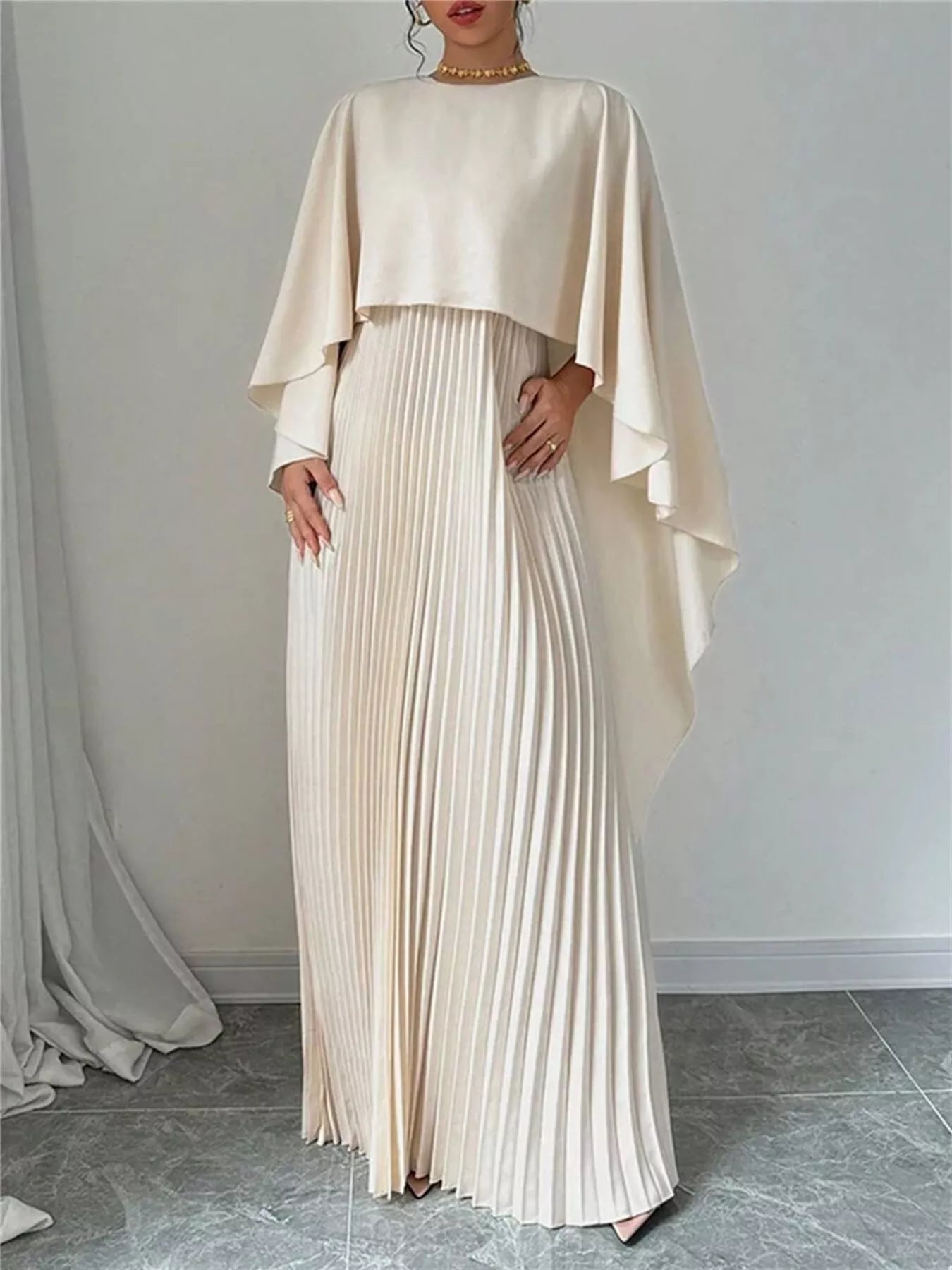 NEWYORKLISTED STYLE COLLECTION: Woman wearing a beige pleated dress with a matching cape against a plain background